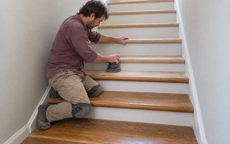 Worker hand-sanding an oak stair tread and its rounded nosing on an enclosed staircase in a two-story Upland house.